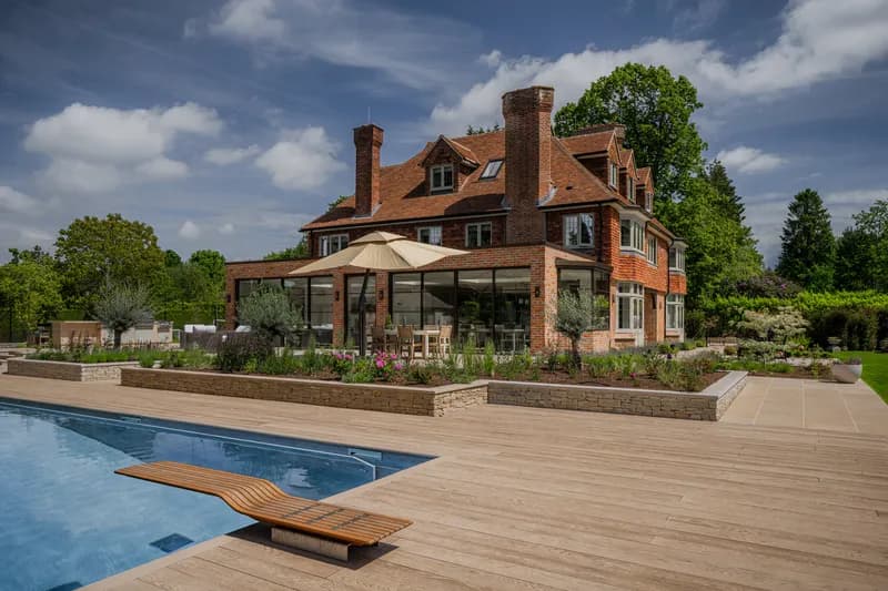 Angled view of an Arts and Crafts red brick house with clay tile roof, tall chimneys, dormers and bay windows, extended with a flat-roofed Crittall-glazed garden room. Raised dry-stone planters in full bloom step down to a heated swimming pool with a curved timber diving board in the foreground, with an outdoor kitchen and seating on the terrace beyond.